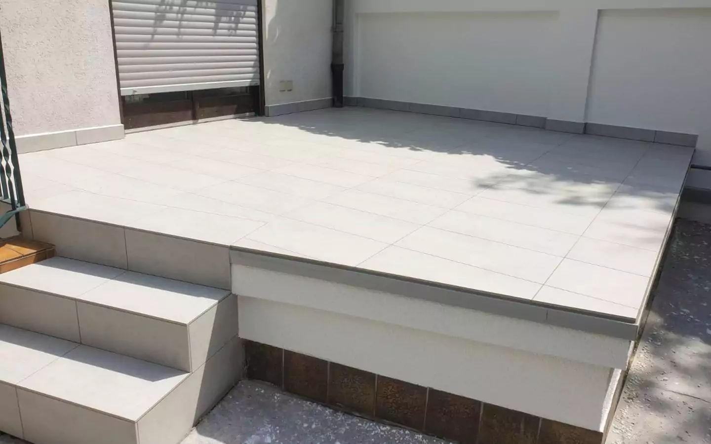 Eine erhöhte Terrasse mit weißen Keramikfliesen, links von Stufen begrenzt und einem teilweise geöffneten Metallladen an der Wand dahinter.