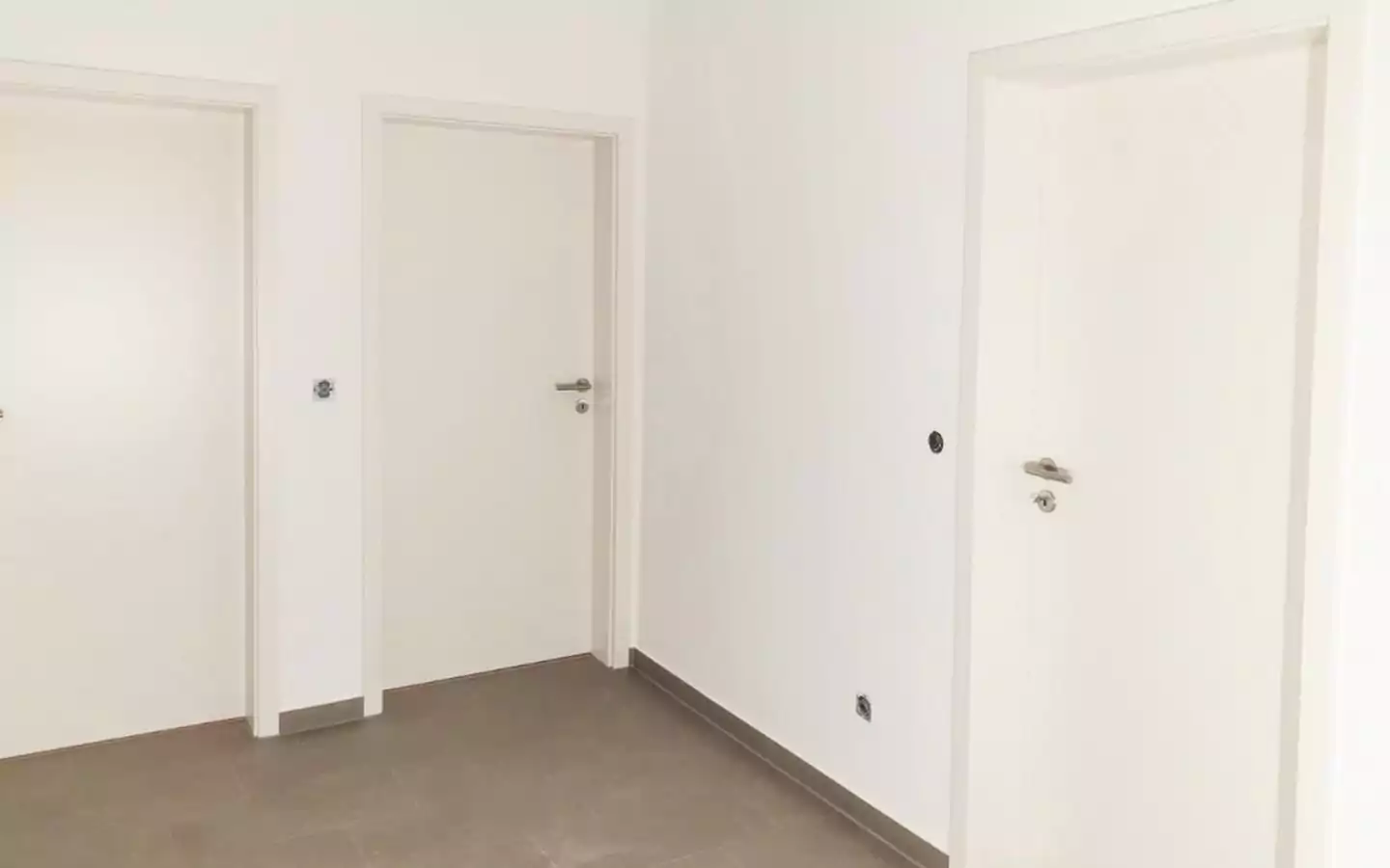 Ein minimalistischer Flur mit weißen Wänden und drei geschlossenen weißen Türen. An einer Wand ist ein Lichtschalter angebracht und der Boden ist mit grauen Fliesen ausgelegt.