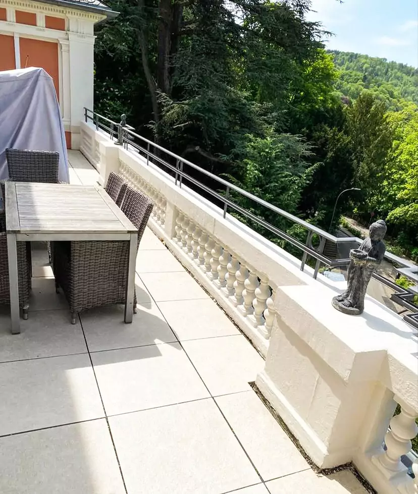 Eine geflieste Außenterrasse mit einem Holztisch, Korbstühlen, weißem Geländer und einer Statue auf dem Sims, umgeben von üppigen grünen Bäumen.