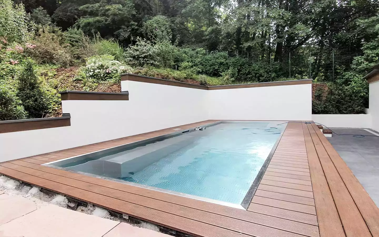 Rechteckiger Swimmingpool mit Holzterrasse, eingebettet zwischen weißen Wänden und üppigem Grün, bietet einen ruhigen Rückzugsort, der an charmante Terrassen erinnert.