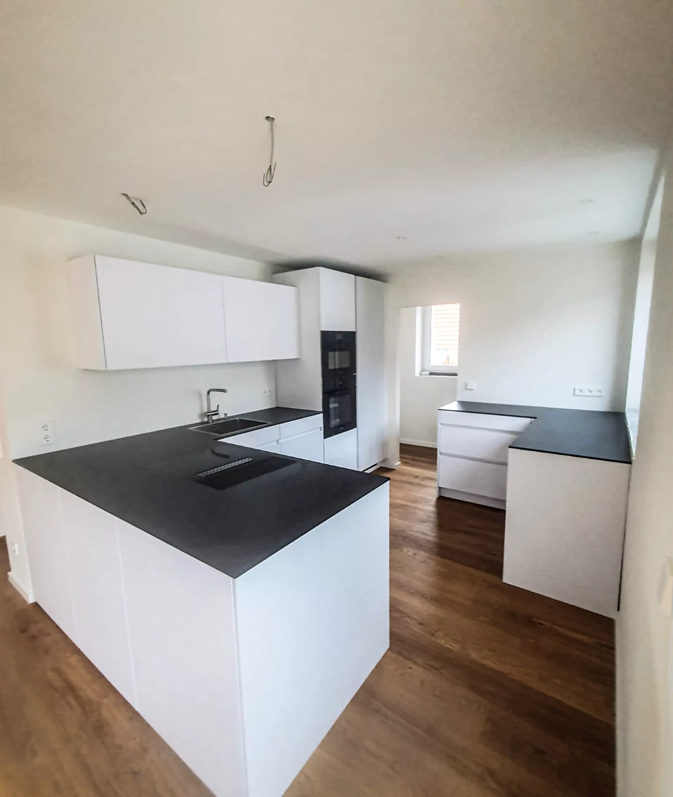 Eine moderne Küche mit weißen Schränken, schwarzen Arbeitsplatten, einem eingebauten Ofen, einer Spüle und einem Fenster. Der Boden ist aus Holz und der Raum ist durch natürliches Licht gut beleuchtet.