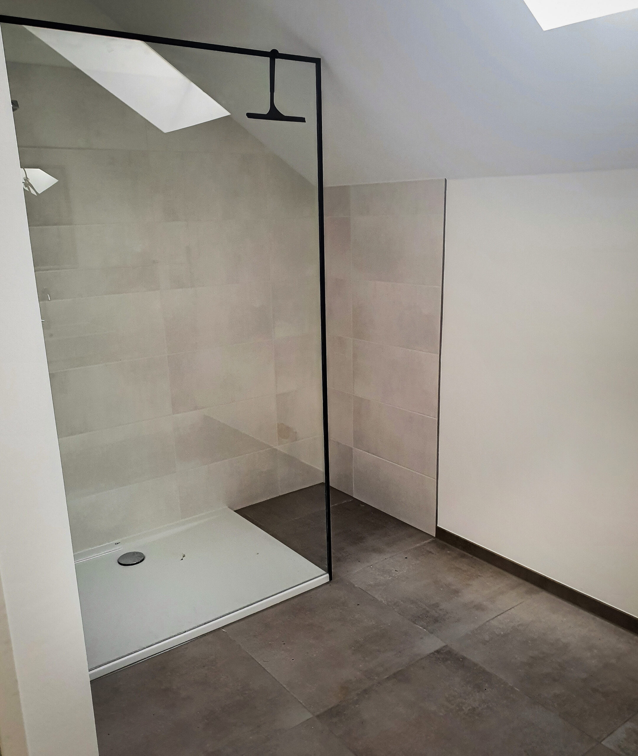 Eine moderne, minimalistische Dusche mit dunkel gefliestem Boden, rahmenloser Glasplatte und Regenduschkopf in einem weiß und beige gefliesten Badezimmer unter einem Oberlicht.