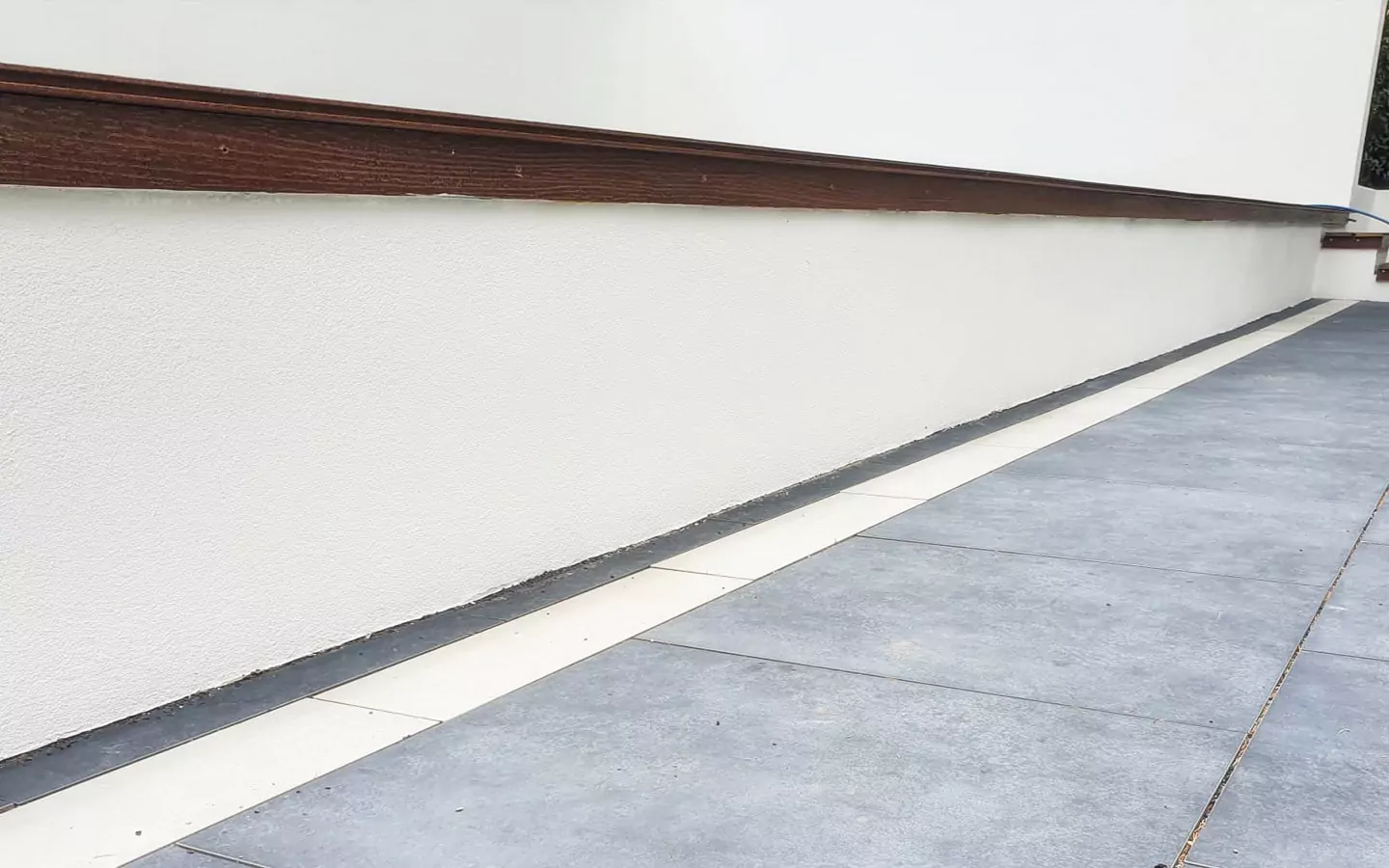 Eine moderne Gebäudeaußenseite mit einer weißen Wand, braunen Holzverkleidungen und einem gepflasterten Gehweg mit großen grauen Fliesen und cremefarbenen Rändern.