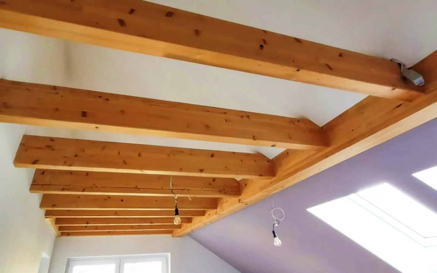 Sichtbare Holzbalken an einer Decke mit zwei Hängelampen und einem schrägen Dach mit Fenstern.