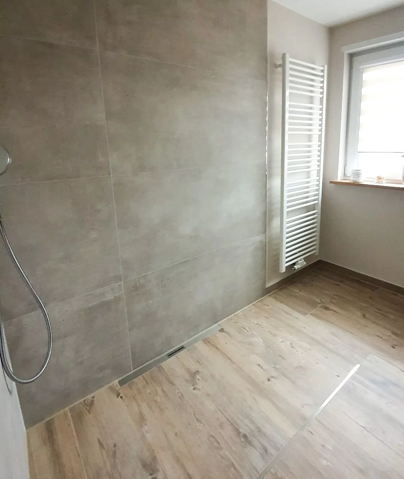 Ein minimalistisches Badezimmer mit Fliesenboden in Holzoptik, großen beigen Wandfliesen, einem Duschkopf auf der linken Seite und einem Fenster mit Heizkörper auf der rechten Seite.