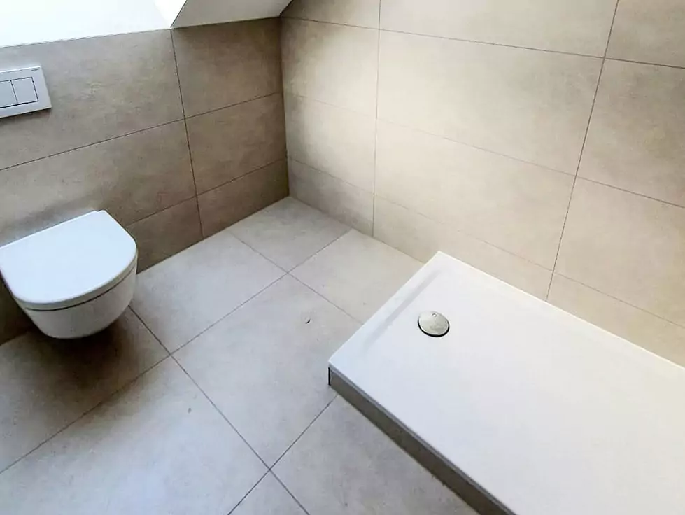 Ein minimalistisches Badezimmer mit beigefarbenen gefliesten Wänden und Böden verfügt über eine wandmontierte Toilette und eine weiße Duschwanne ohne Wände oder Vorhang.