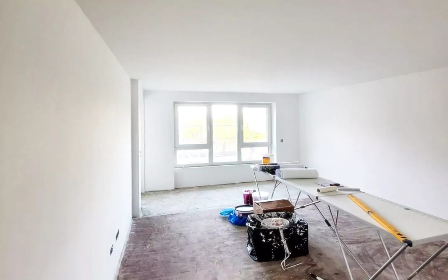 Ein im Renovierungsstadium befindlicher Raum mit weißen Wänden, großen Fenstern und verschiedenen Baumaterialien und Werkzeugen, die auf dem Boden und einem Tisch verstreut sind.