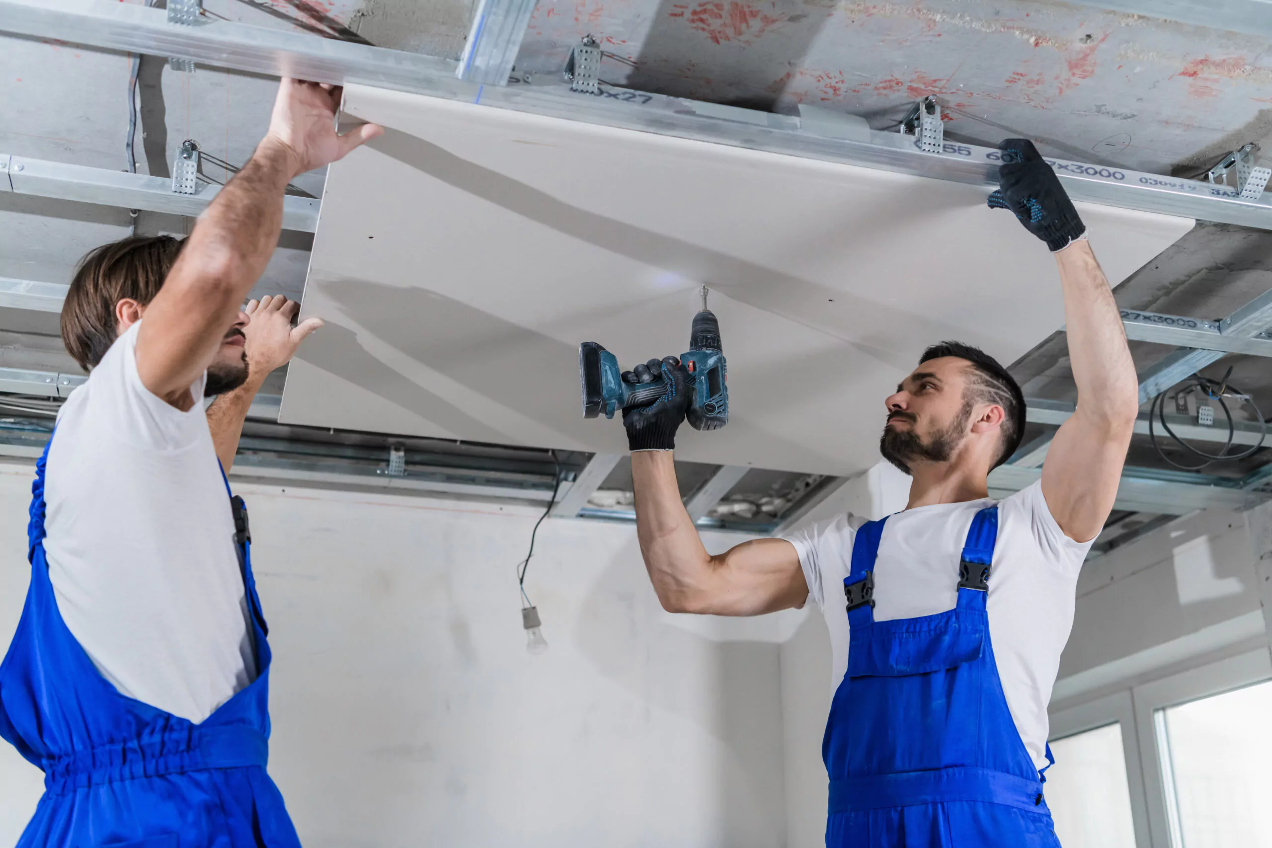 Zwei Bauarbeiter in blauen Overalls montieren mit einer Bohrmaschine eine Gipskartonplatte an der Decke.
