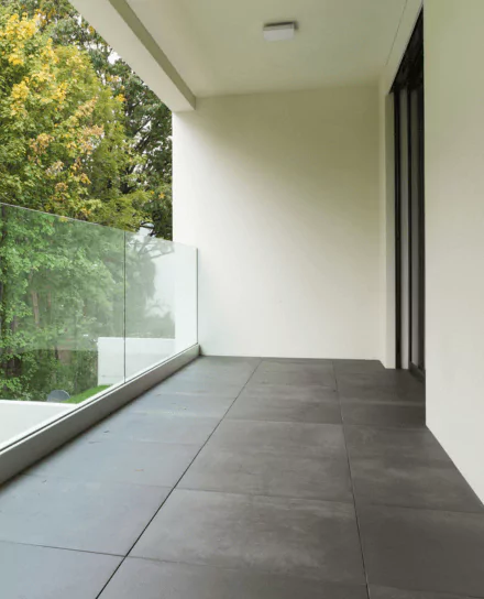 Ein minimalistischer Balkon mit grauen Bodenfliesen, einem Glasgeländer und Blick auf grüne Bäume.