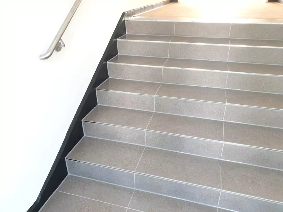 Eine gut beleuchtete Treppe mit grau gefliesten Stufen und einem metallischen Handlauf auf der linken Seite führt nach oben.