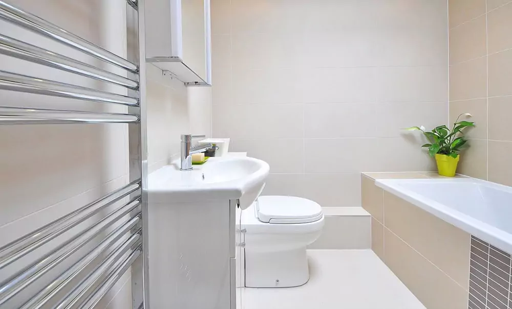 Ein modernes Badezimmer mit weißem Waschbecken, Toilette, wandmontiertem Handtuchheizkörper, Badewanne und einer grünen Topfpflanze auf dem Wannenrand. Die Wände sind in Beige und Weiß gefliest.