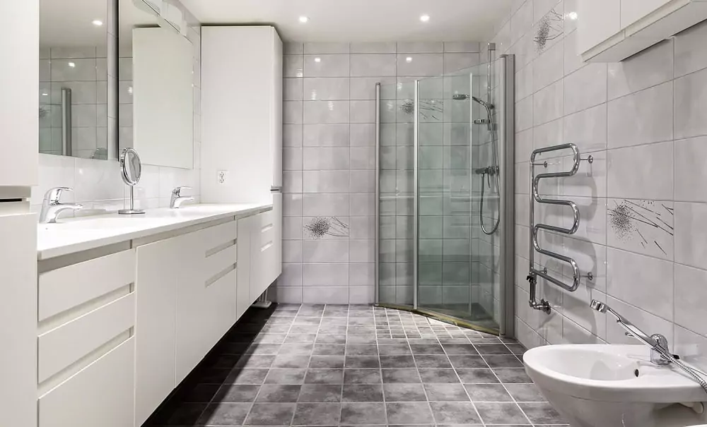 Modernes Badezimmer mit weißen Schränken, Doppelwaschbecken, verglaster Dusche, wandmontiertem Handtuchwärmer und Bidet. Das Zimmer hat grau geflieste Wände und einen grau gefliesten Boden, was die Qualität der Badsanierung unterstreicht.