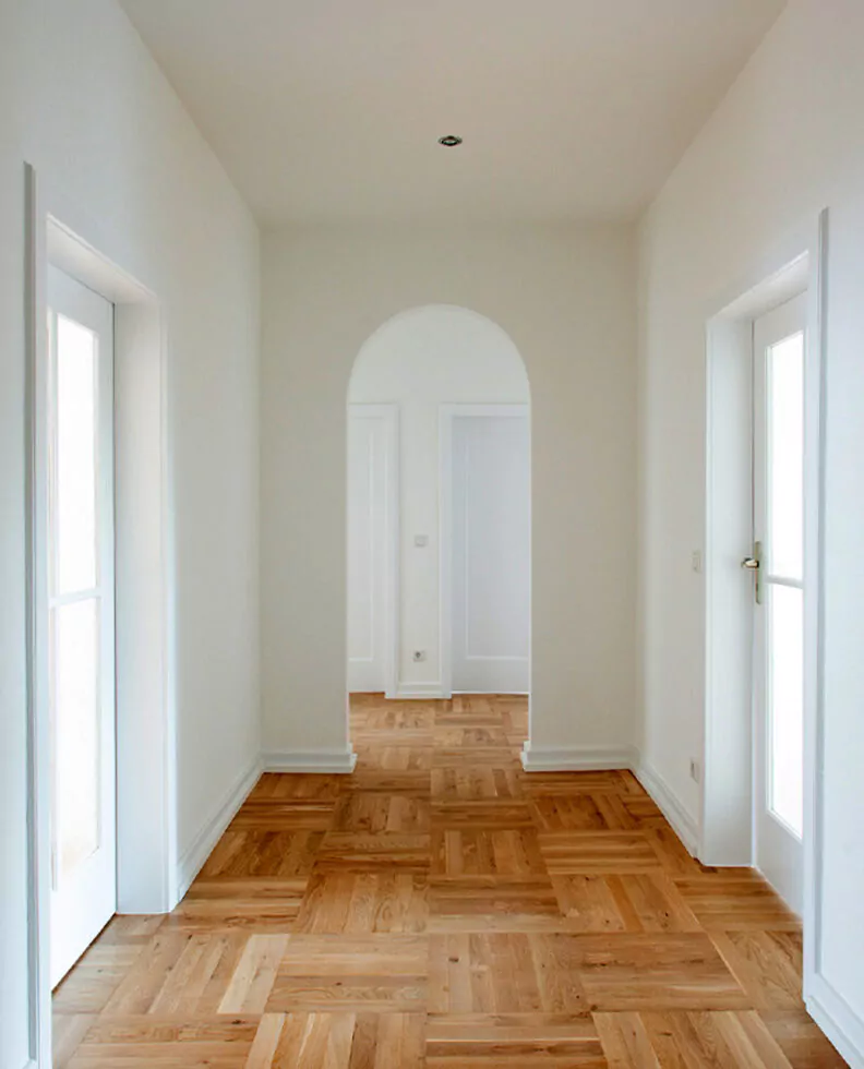 Ein Flur mit Holzparkettboden, das Ergebnis sorgfältiger Bodenlegearbeiten, weist weiße Wände und Türrahmen auf. Am Ende des Flurs ist eine gewölbte Tür zu sehen.