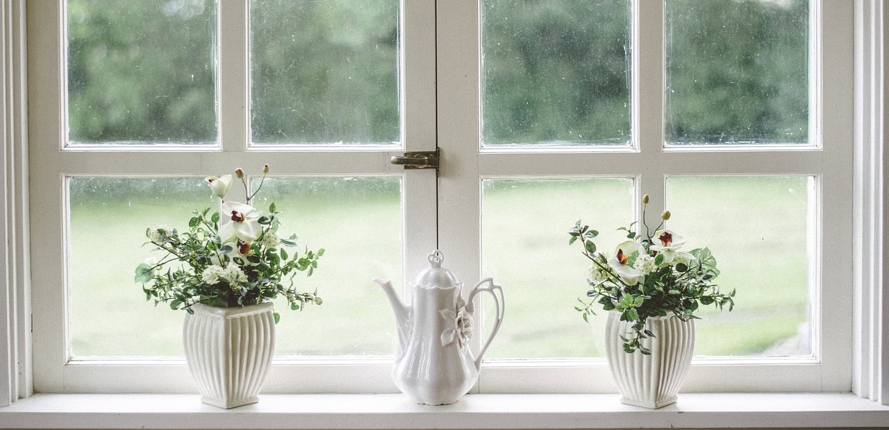 Zwei weiße Blumenvasen mit Grünpflanzen und Blumen stehen auf beiden Seiten einer weißen Teekanne auf einem Fensterbrett, mit Blick ins Grüne draußen.