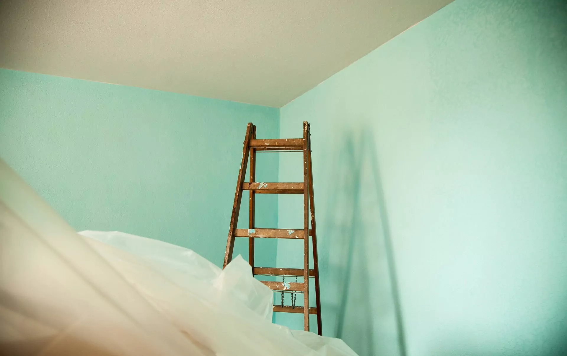 An einer hellblauen Wand neben einer Decke steht eine Holzleiter, im Vordergrund ist eine Plastikfolie über den Möbeln angebracht.