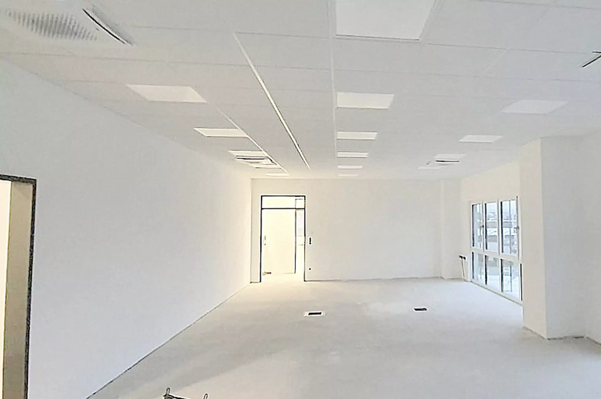 Ein leerer Büroraum mit weißen Wänden, einer weißen Decke mit Einbauleuchten, großen Fenstern auf der rechten Seite und einer Tür am anderen Ende. Der Boden ist unmöbliert und kahl.