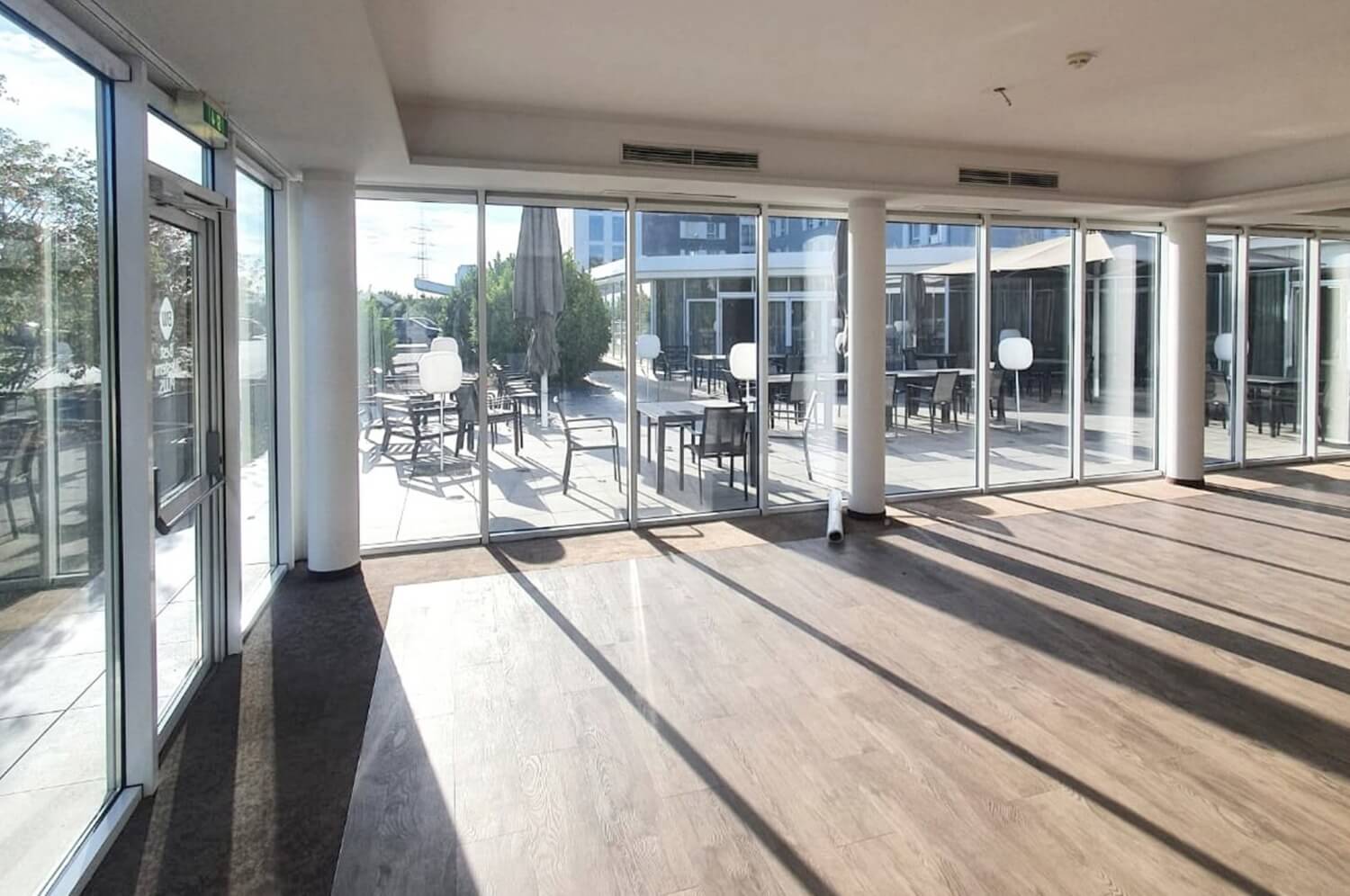 Geräumiger Innenbereich mit Holzboden und großen Glastüren, die auf eine Außenterrasse mit Tischen und Stühlen führen. Sonnenlicht wirft Schatten auf den Boden und schafft eine warme Atmosphäre in diesem charmanten Hotelrestaurant nach einem kürzlichen Umbau.