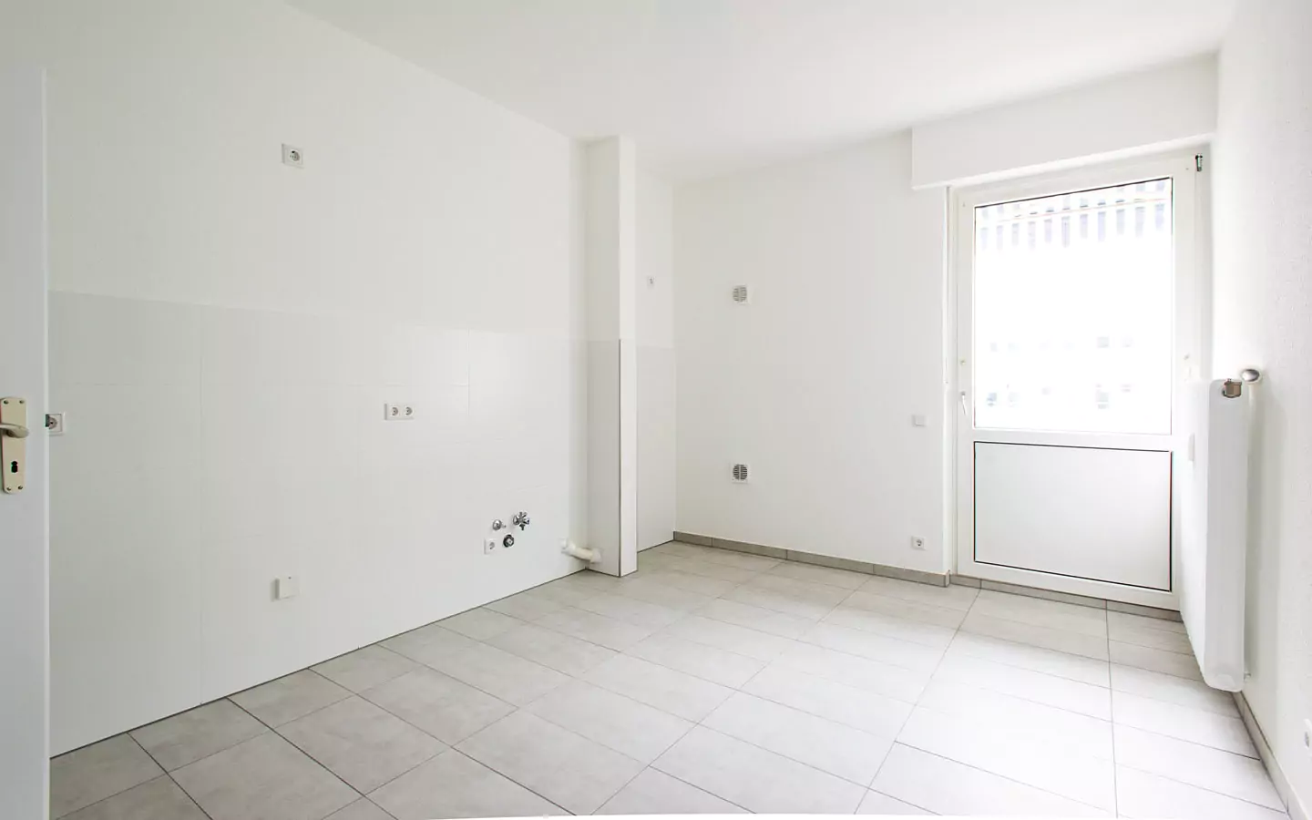 Ein leerer, weiß gefliester Raum in einer Altbauwohnung mit geschlossener Tür nach draußen, sichtbaren Sanitär- und Steckdosen sowie einem einzelnen Heizkörper an der rechten Wand, der auf seine Komplettsanierung wartet.