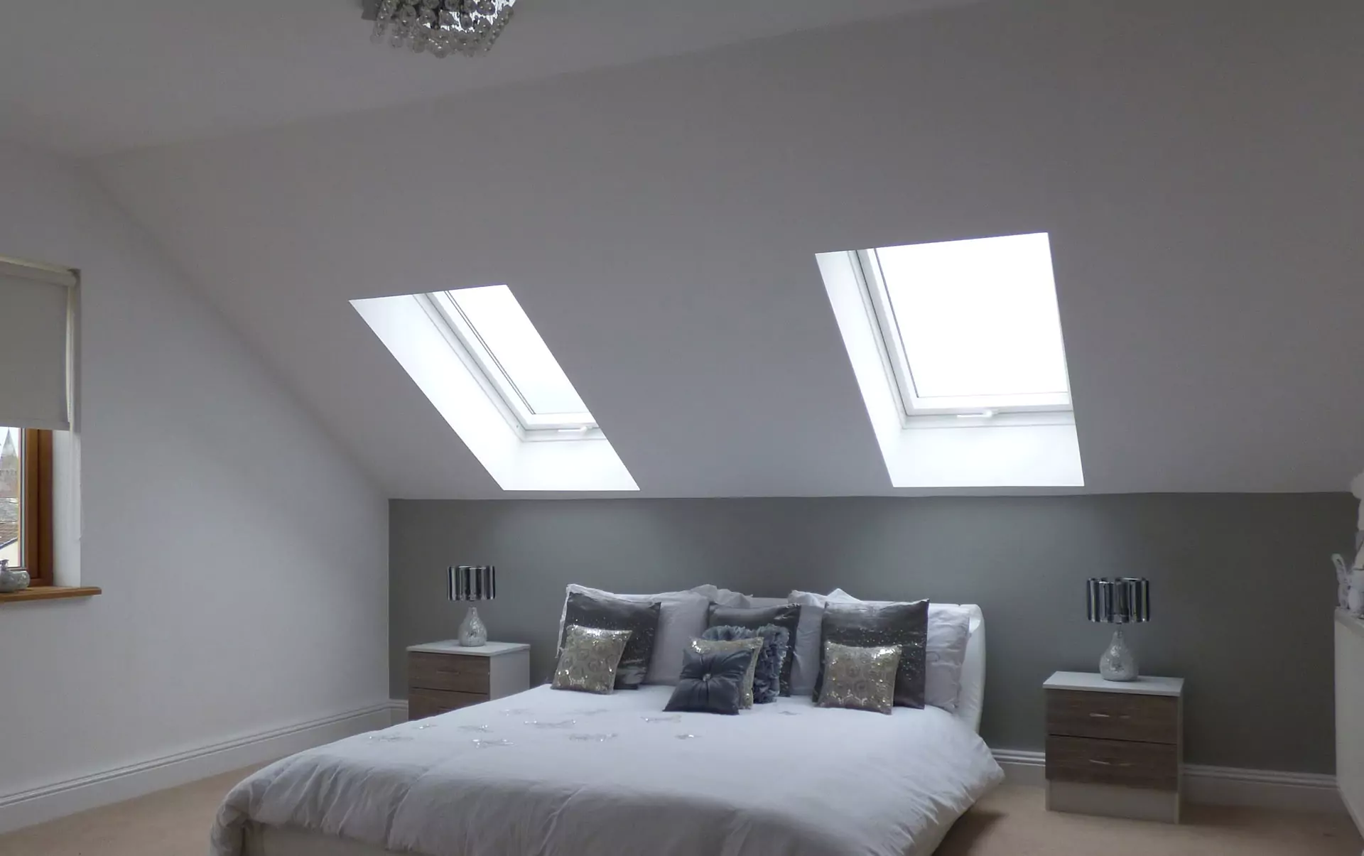 Ein Schlafzimmer mit einem Doppelbett, grauen und silbernen Kissen, zwei Oberlichtern, einer hellgrauen Akzentwand, Nachttischen mit Tischlampen und einem Kronleuchter. An der linken Seitenwand befindet sich ein Fenster mit Jalousie.