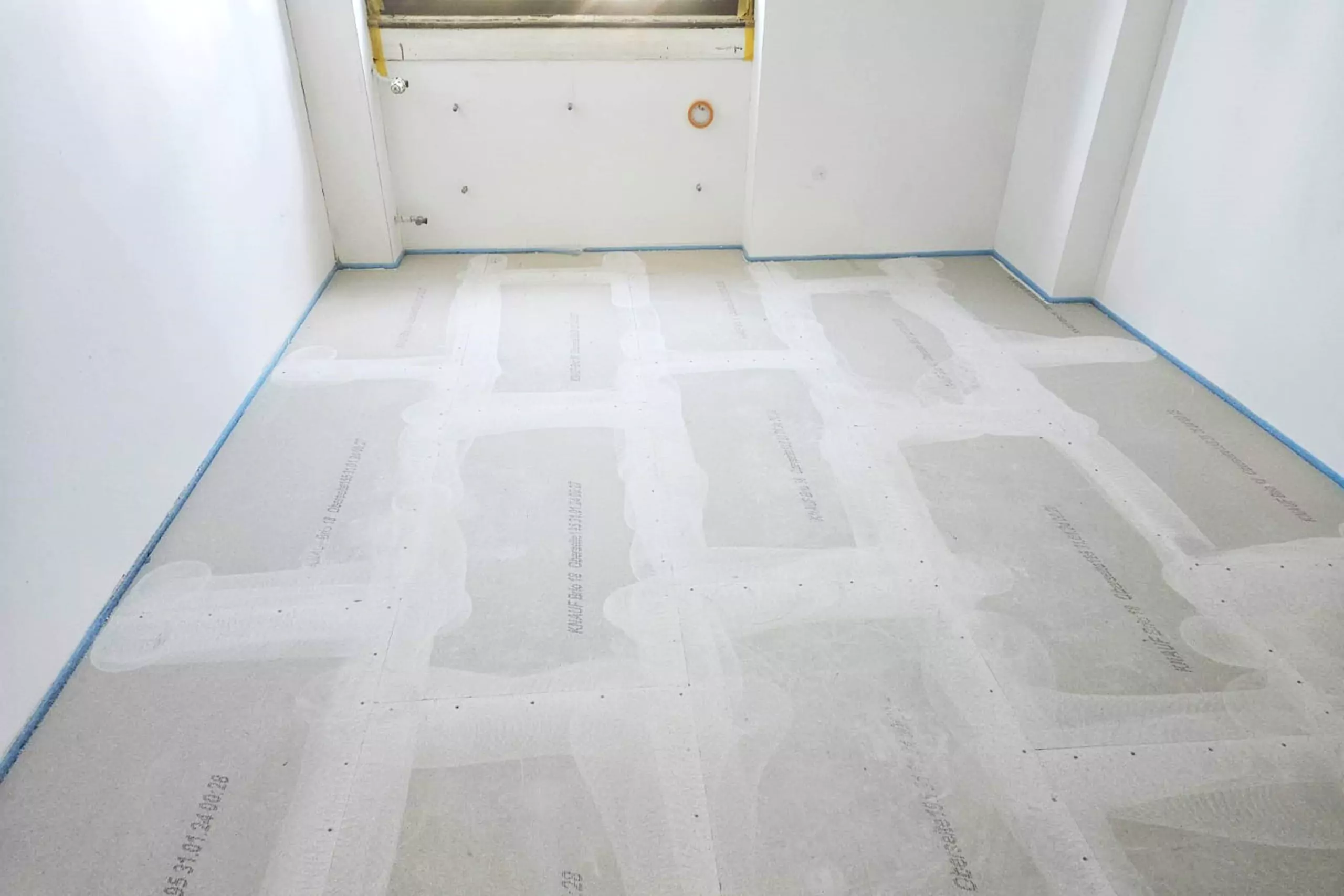 Ein Raum im Umbau mit ungestrichenen Wänden und einem für weitere Arbeiten vorbereiteten Boden. Der Bodenbelag scheint unfertig zu sein und an den Rändern sind einige Baumaterialien sichtbar.